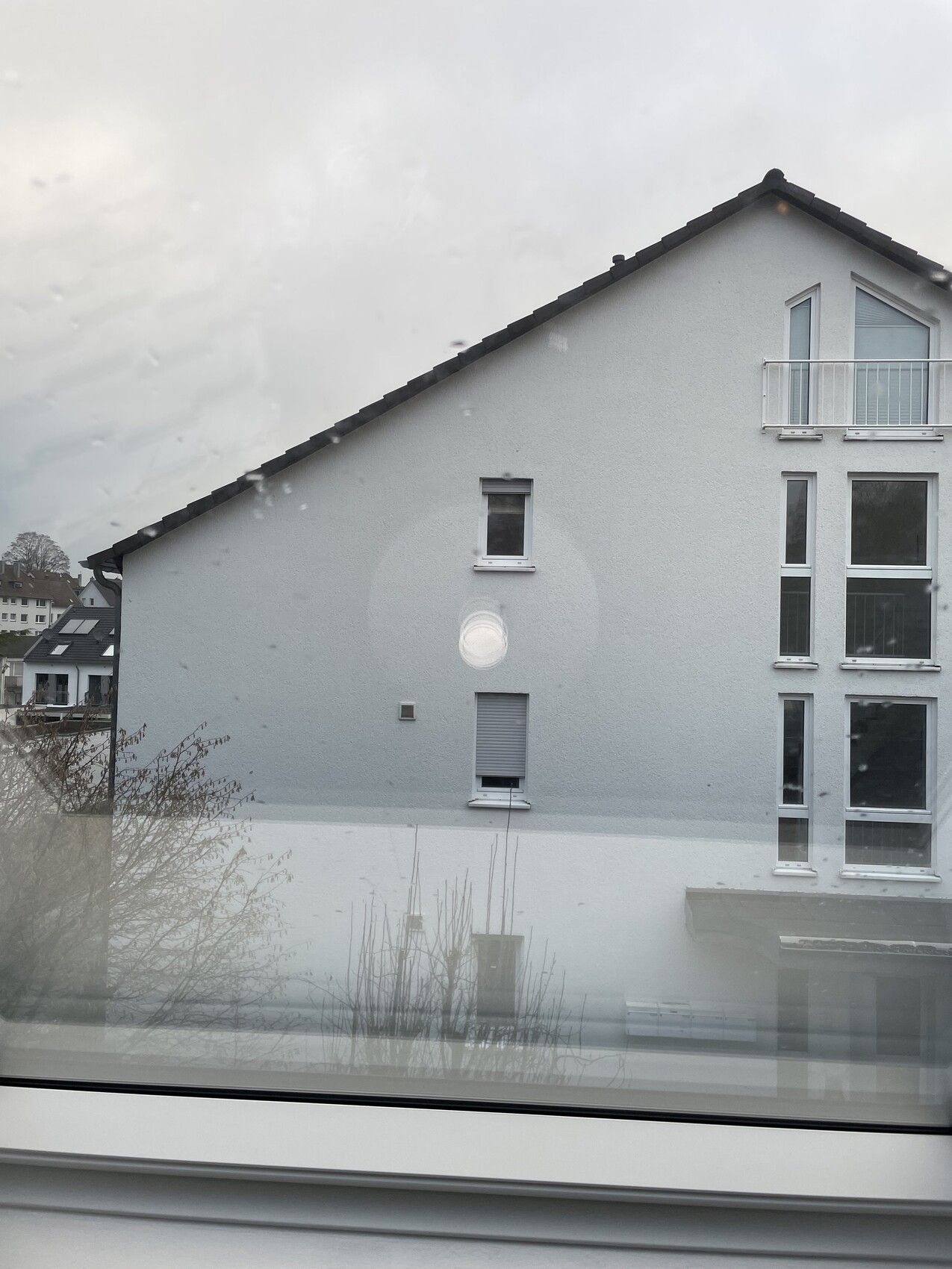 Weißes Einfamilienhaus mit Satteldach, großen Fenstern und Balkon, Sicht durch regennasse Scheibe.