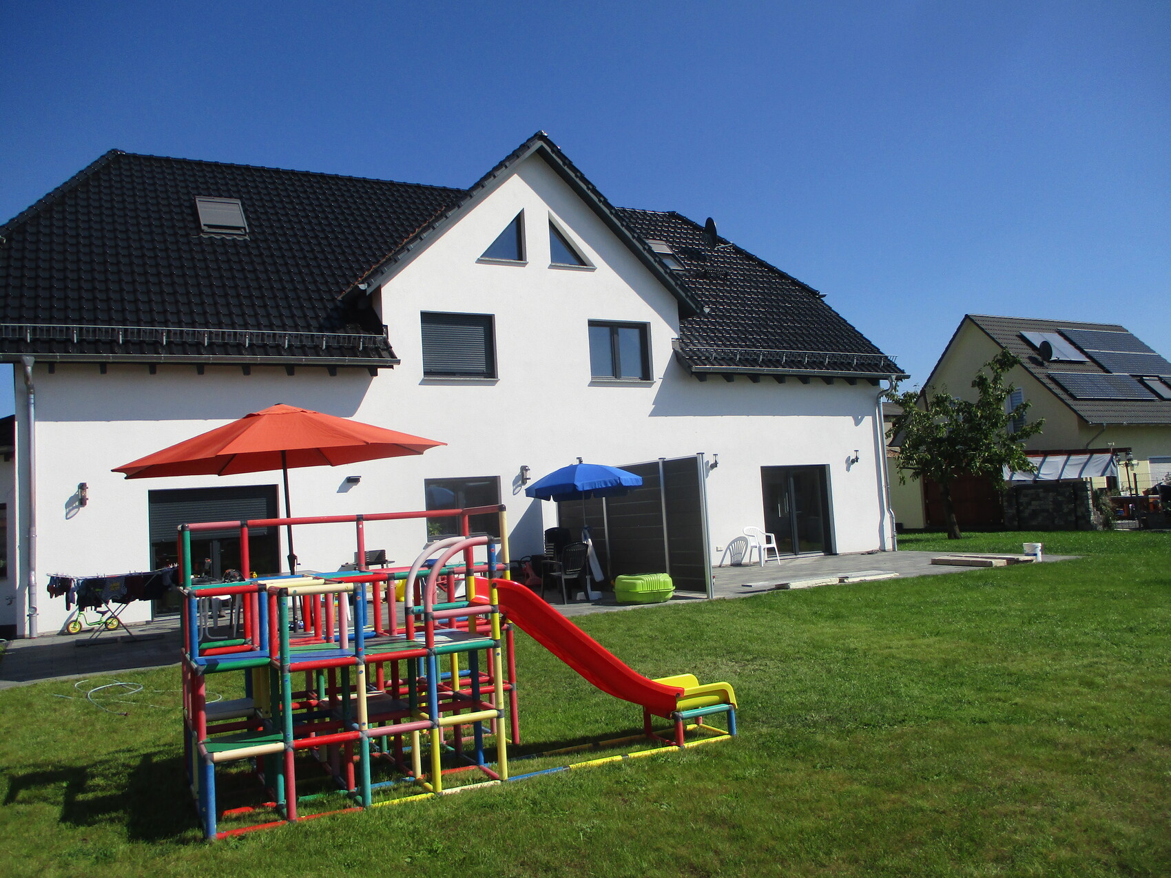 Garten hinter weißem Einfamilienhaus mit farbigem Klettergerüst, roter Rutsche und Sonnenschirmen.