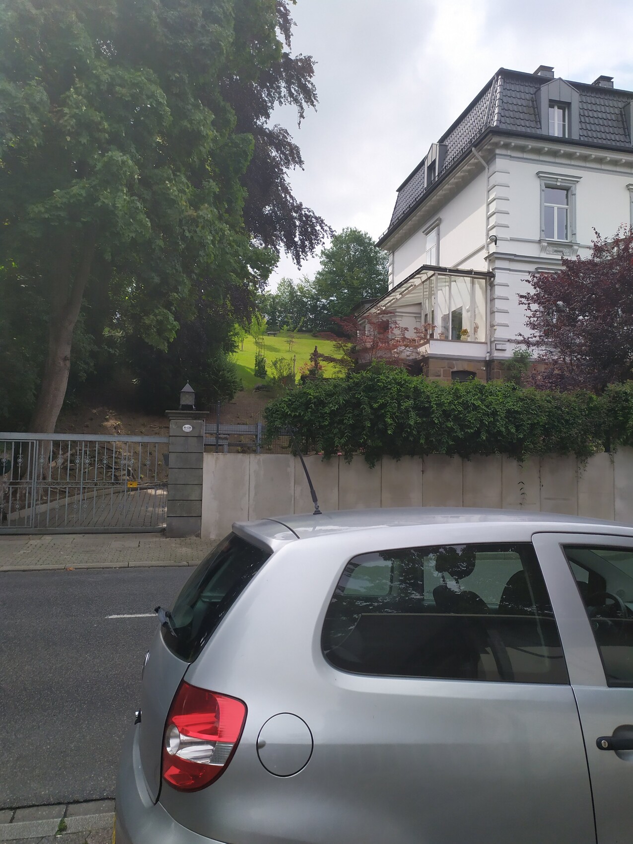 Silbernes Auto vor weißem Villengebäude mit Glasveranda und Garten.