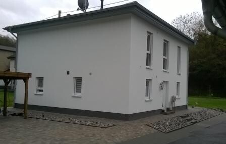 Kleines weißes Einfamilienhaus mit zwei Stockwerken, flacher Dachkante und Satellitenschüssel.