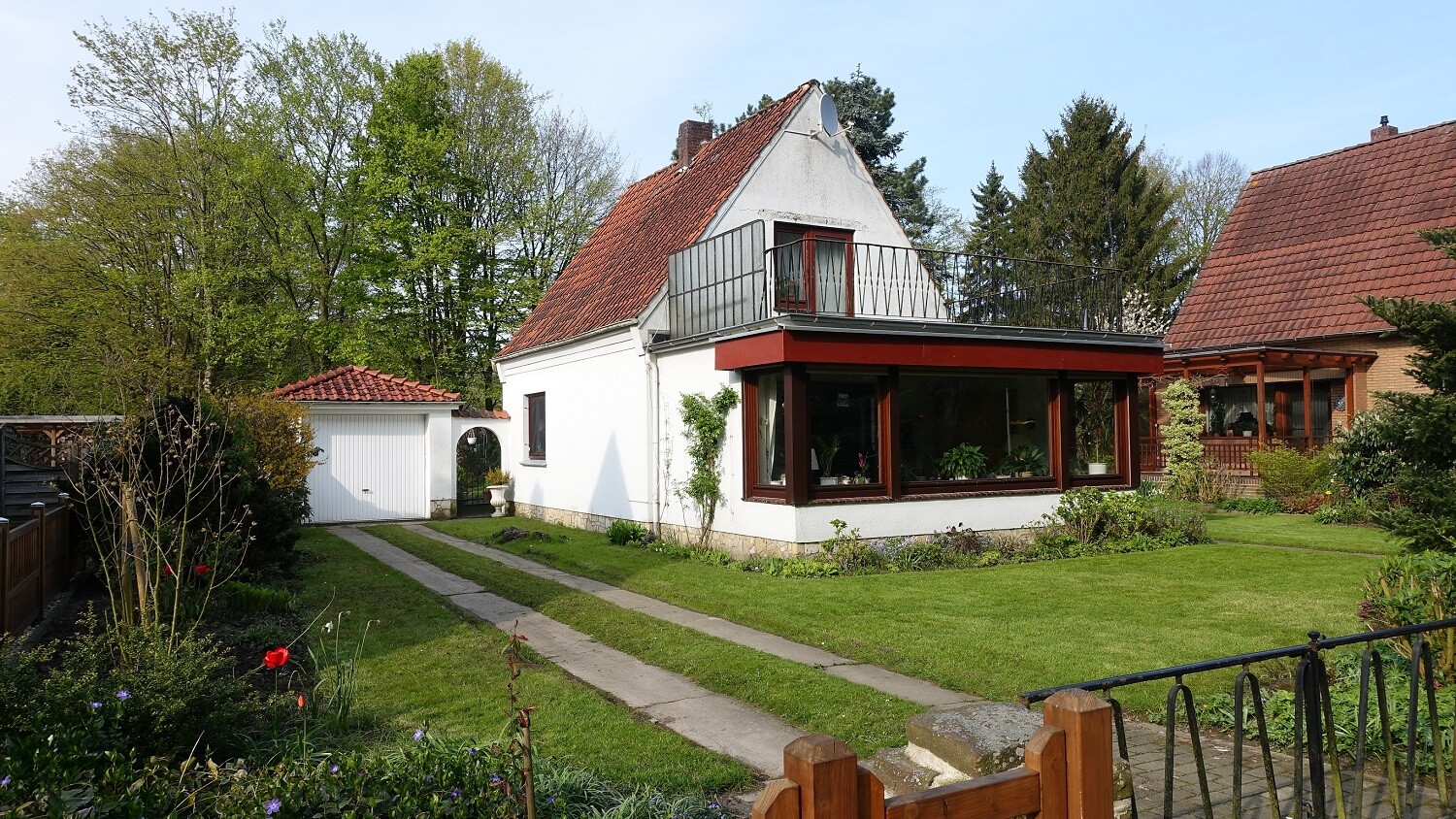 Weißes Haus mit rotem Ziegeldach, Glasveranda und Balkon, gepflegter Garten und Garage.