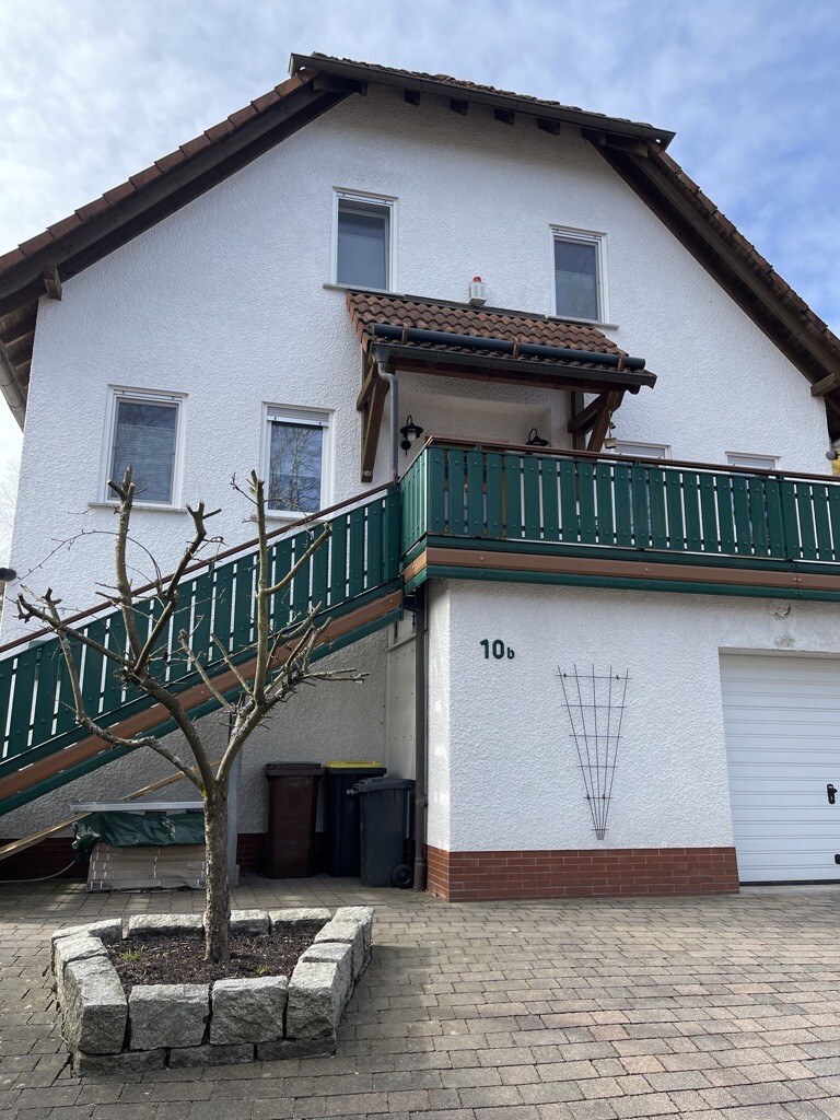 Weiße Wohnhausfassade mit grünem Balkon, Außentreppe, Baum im Pflanzbeet und Mülltonnen davor.