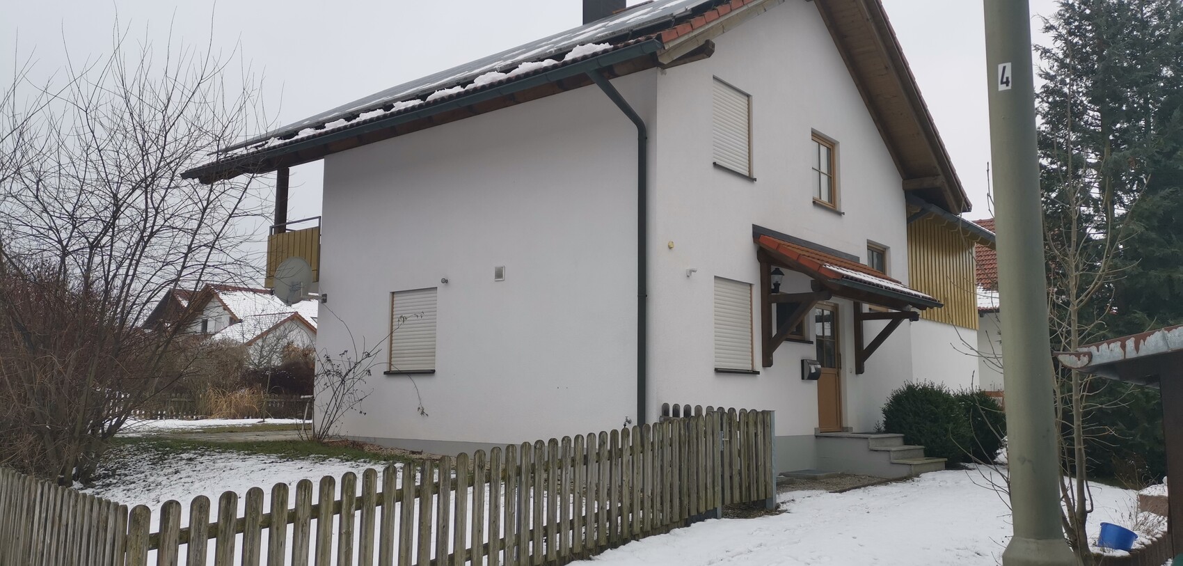 Weißes zweistöckiges Haus mit Vordach, verschneitem Hof, Holzzaun und Satellitenschüssel.