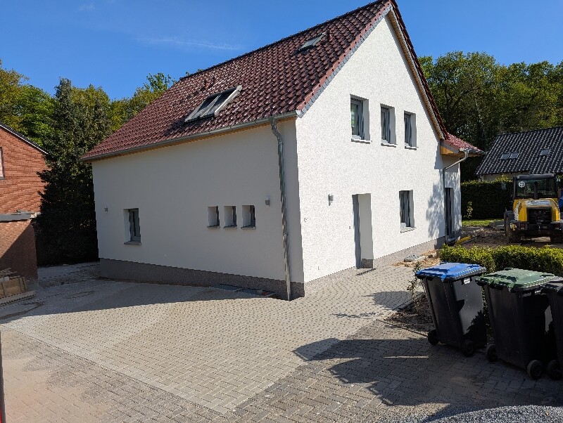 Weißes, zweistöckiges Haus mit rotem Ziegeldach, gepflasterter Hof und Mülltonnen davor.