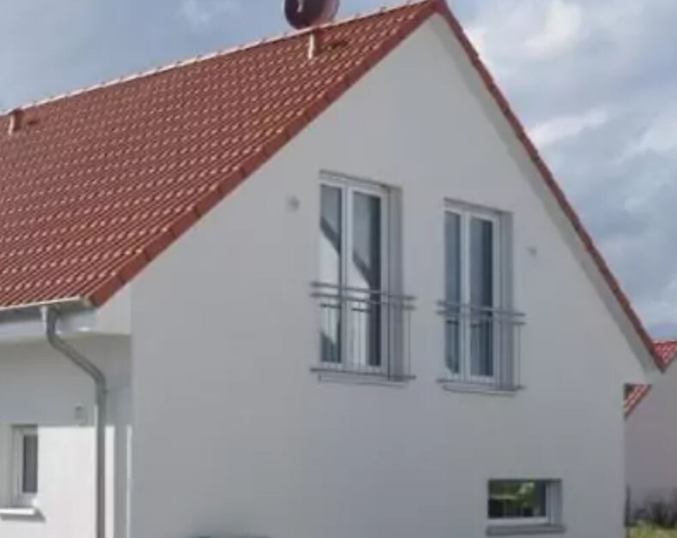 Weißes Haus mit rotem Ziegeldach, zwei obere Fenster mit Geländern.
