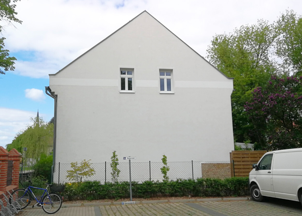Helles Haus mit spitzem Giebeldach, zwei Fenster, Zaun, Fahrrad links, Van rechts, Bäume dahinter