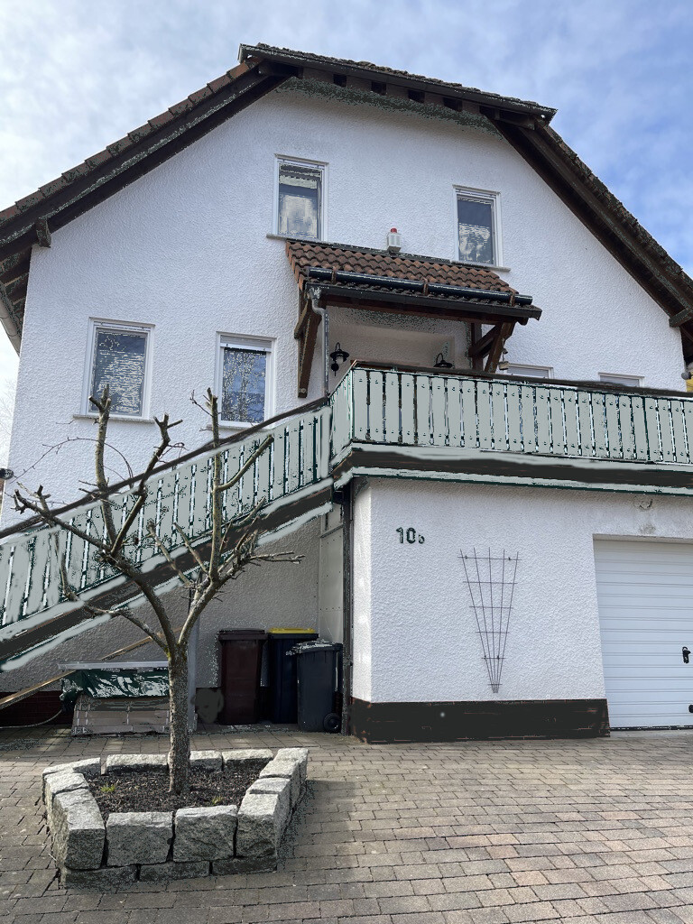 Außenansicht eines weißen Hauses mit Außentreppe, Balkon, Baum im Pflanzbeet und Mülltonnen.