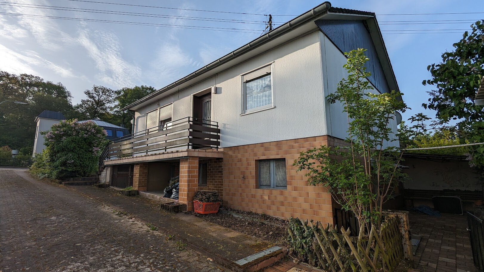 Zweistöckiges Haus mit weißer Fassade, Klinkerunterbau, Holz-Balkon und Baum davor.