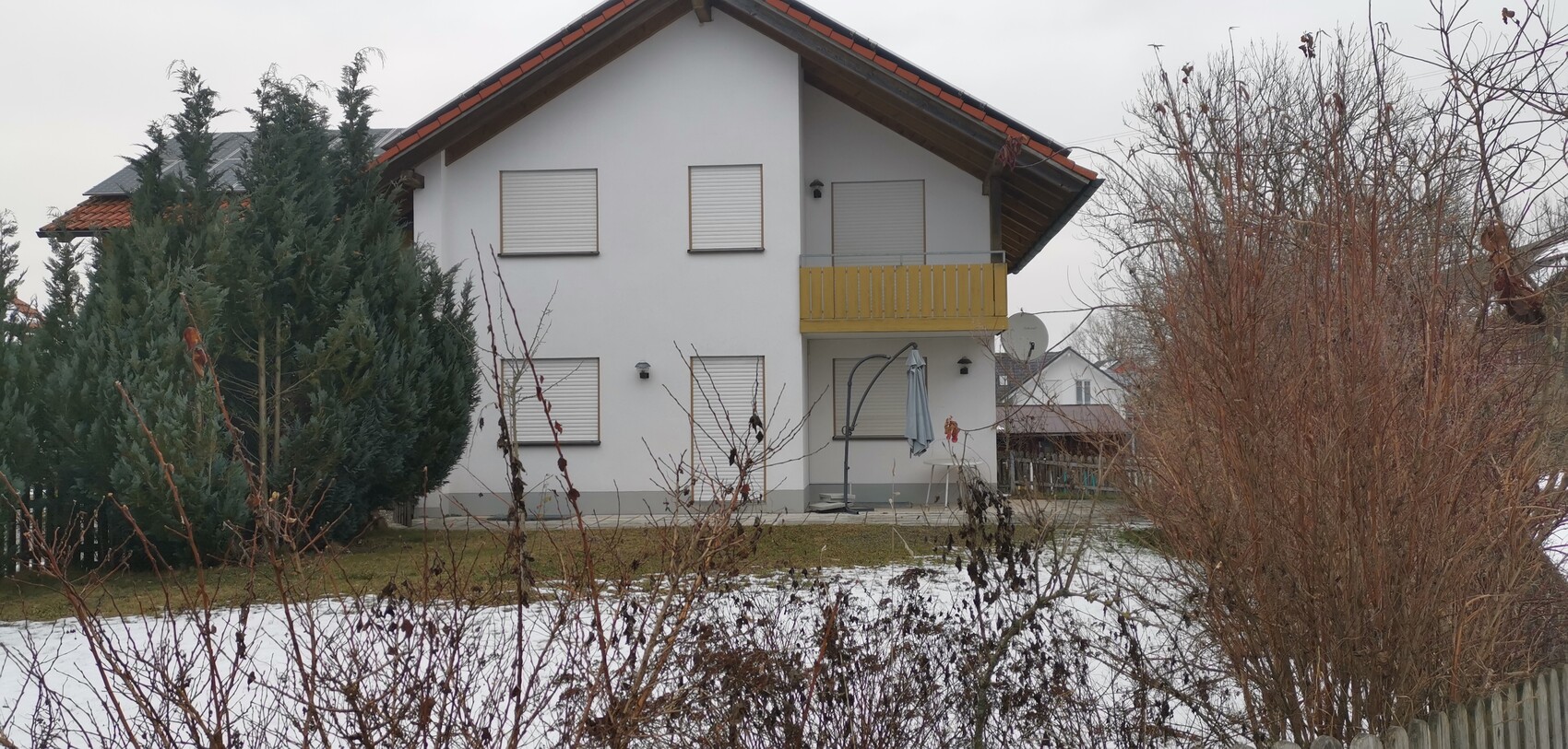 Zweistöckiges weißes Haus mit gelbem Balkon, geschlossenen Rollläden, Schnee und Bäumen.
