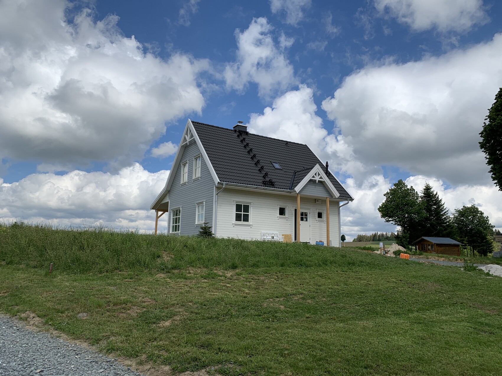 Weißes Einfamilienhaus mit grauem Satteldach auf grünem Hügel vor Wolkenhimmel.