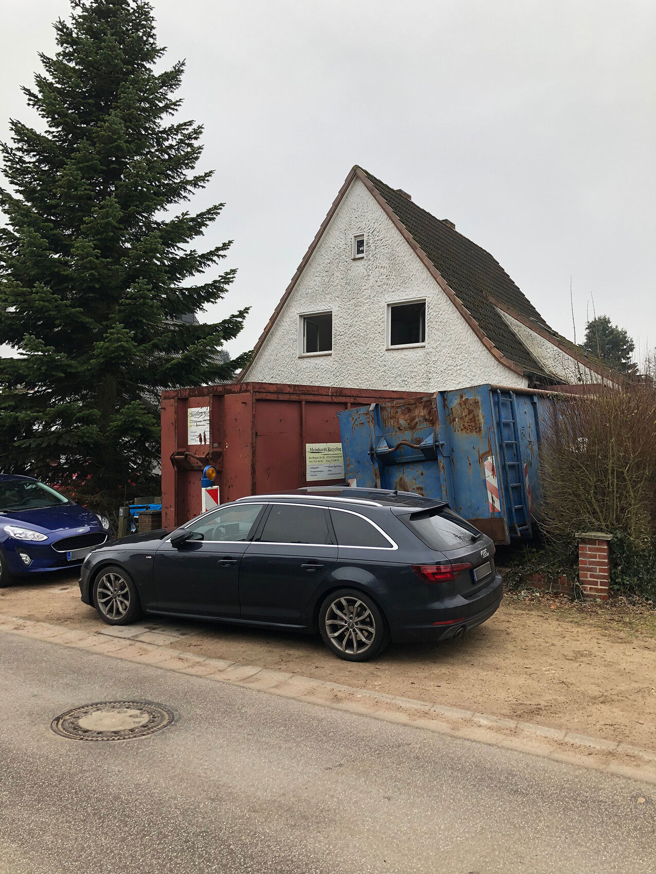 Weißes Spitzhaus mit zwei Fenstern; davor rote und blaue Müllcontainer, grauer Kombi, links Baum.