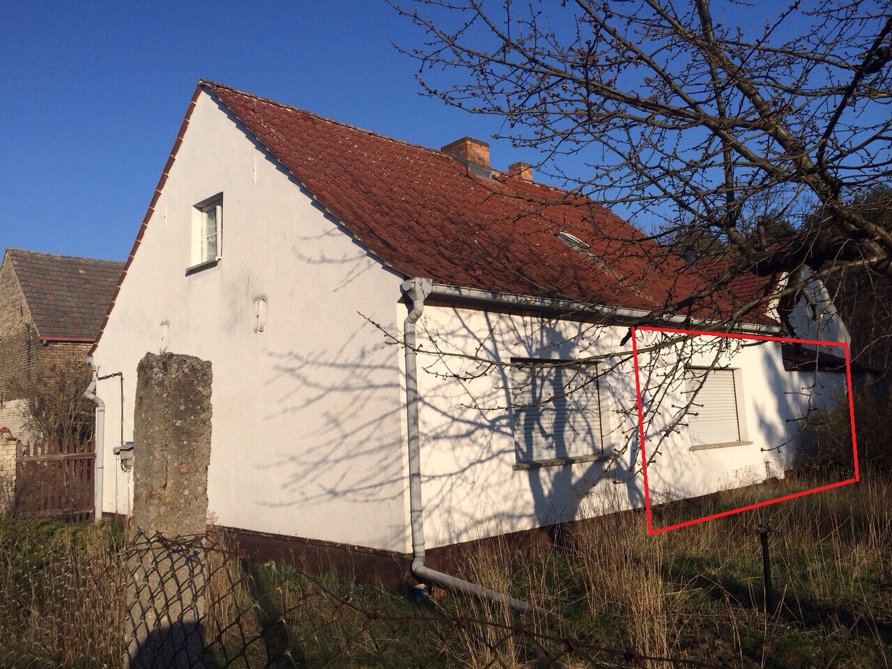 Weißes Haus mit rotem Dach; rechte Wandteil im roten Rahmen, davor Baumzweige, blauer Himmel.