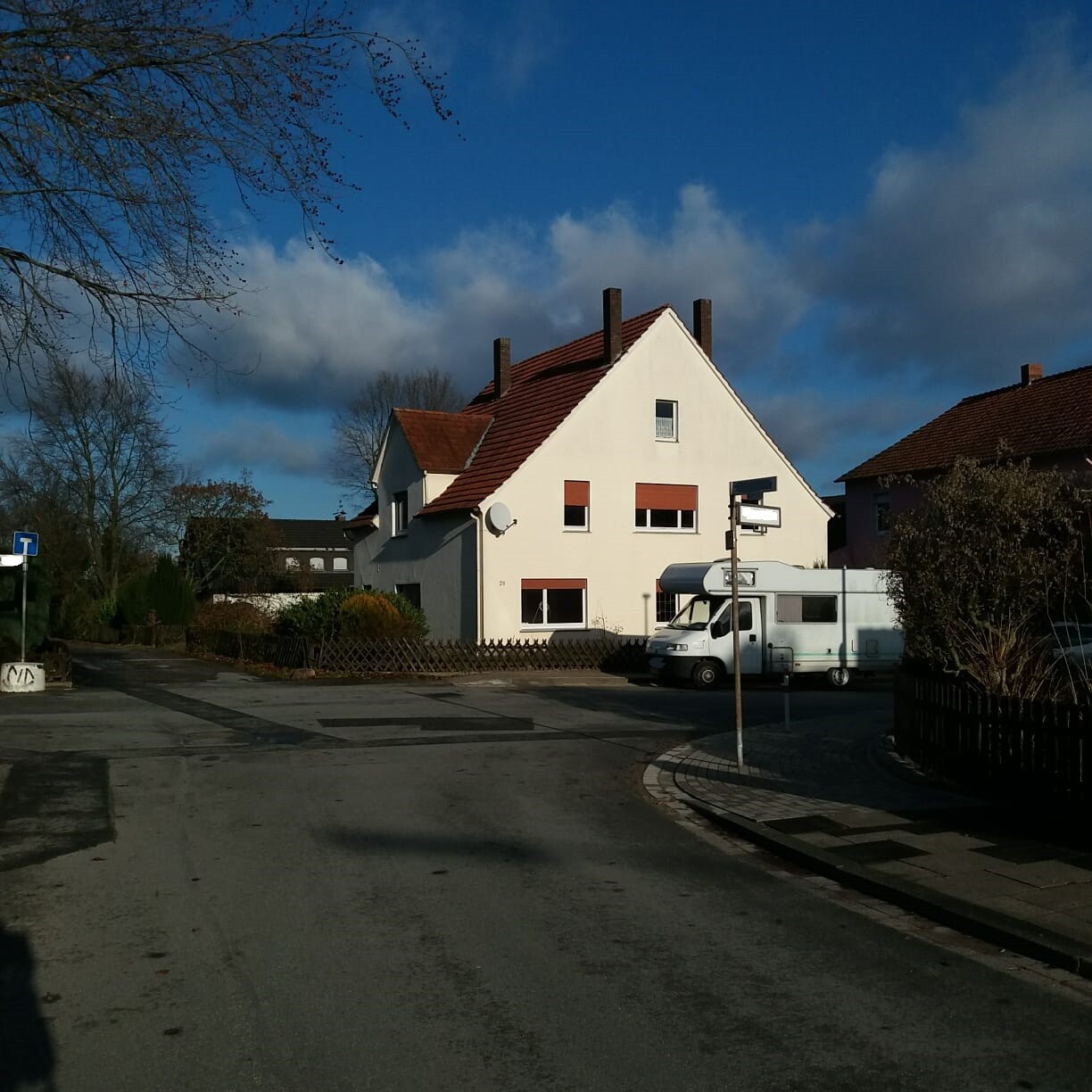Weißes Einfamilienhaus mit rotem Dach, davor ein Wohnmobil, Straßenecke unter blauem Himmel.