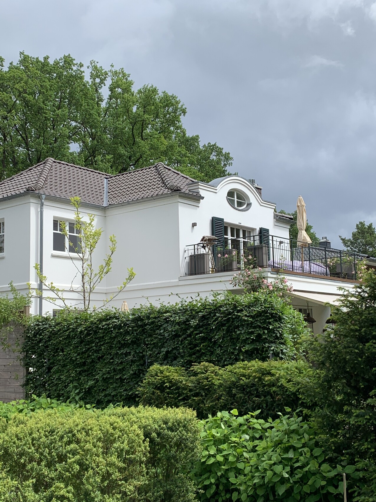 Weißes zweistöckiges Haus mit Balkon, Terrasse, umgeben von Hecken und Bäumen, wolkigem Himmel.