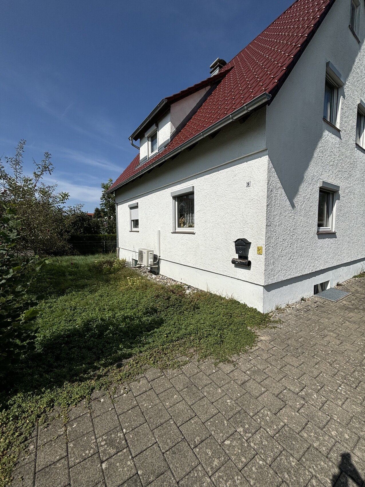 Weißes Einfamilienhaus mit rotem Ziegeldach, Garten links und gepflastertem Weg vor dem Haus.