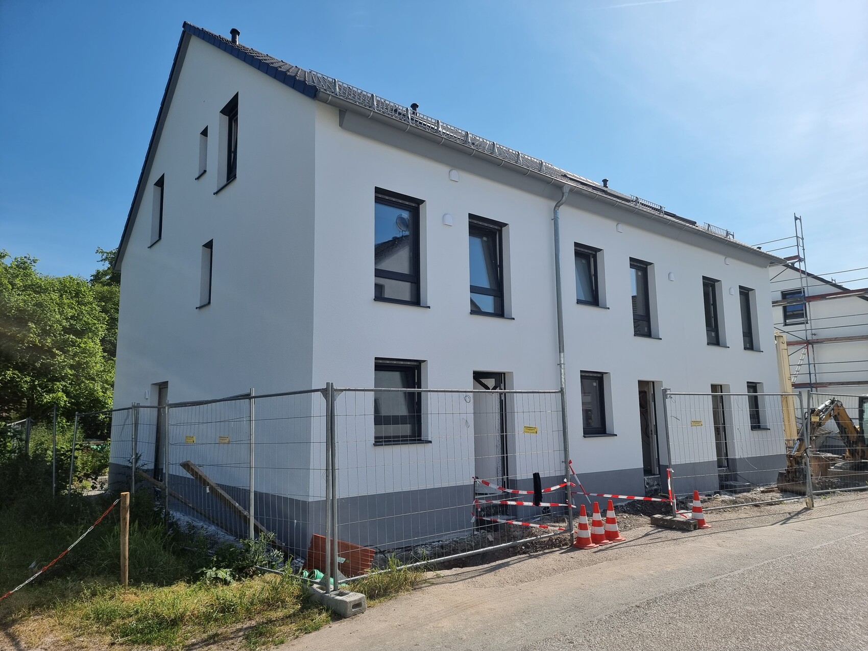 Weißes, zweistöckiges Neubauhaus hinter Bauzaun; Baukegel und Gerüst sichtbar.