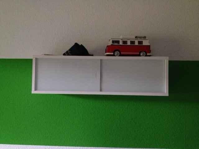 Weißes Wandregal mit rotem Spielzeugbus an grüner Wand.