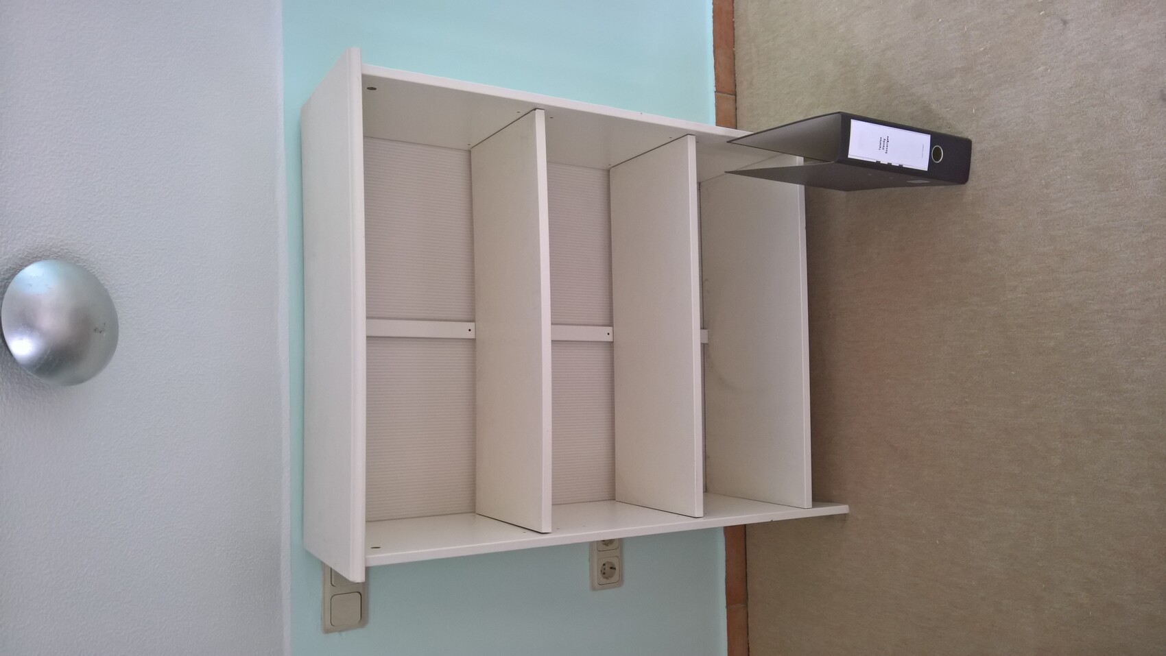 Weißes Regal im Wohnzimmer vor hellblauer Wand, Ordner daneben, beige Boden.