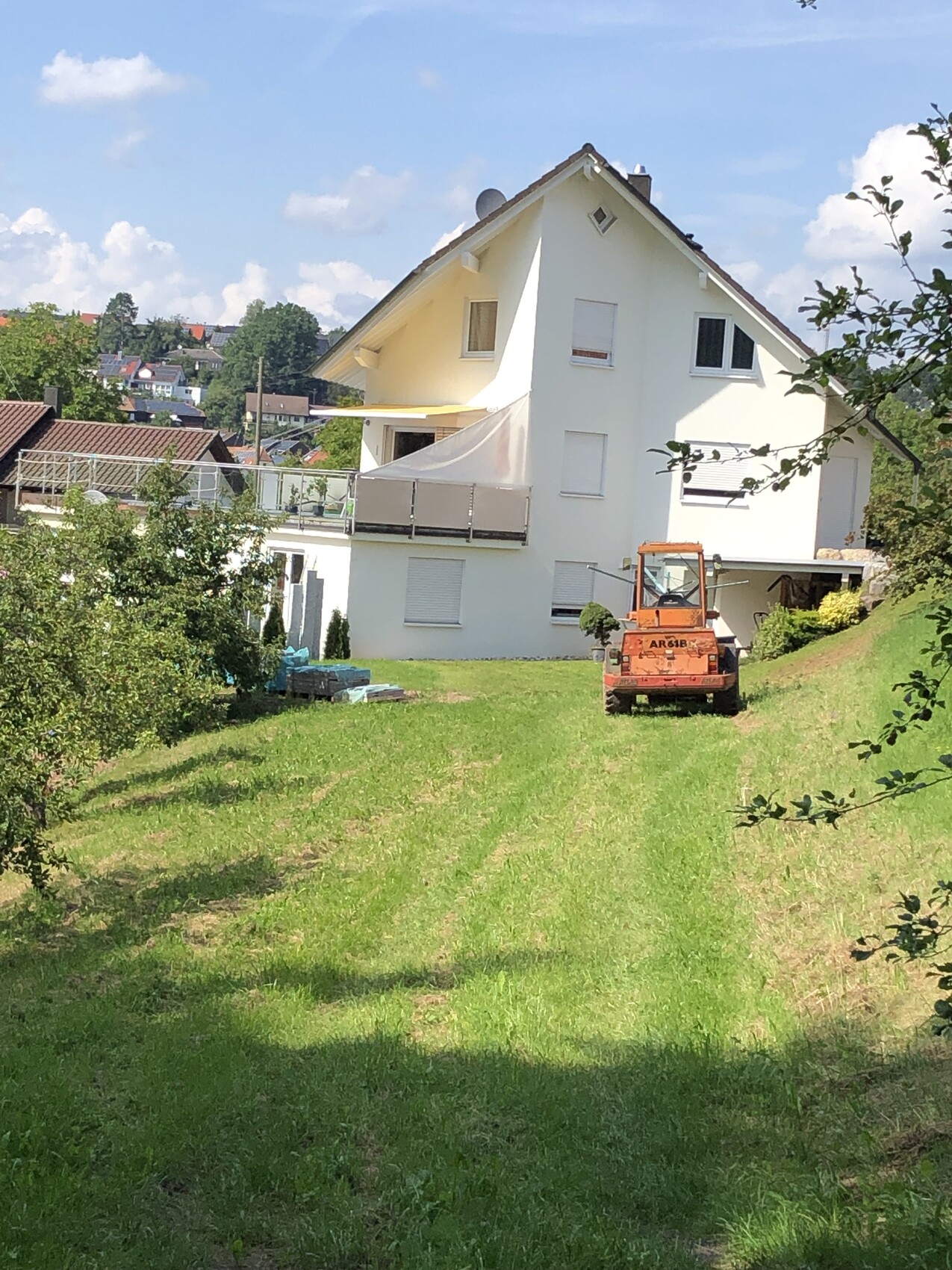 Weiße mehrstöckige Villa auf Hang mit Terrasse; orange Baumaschine im Garten, grüne Wiese.