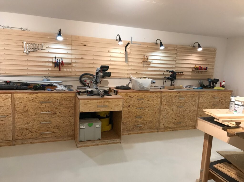 Werkstatt mit Holzwerkbank, OSB-Schränken, Kreissäge, Bohrmaschine und Werkzeugen an der Wand.