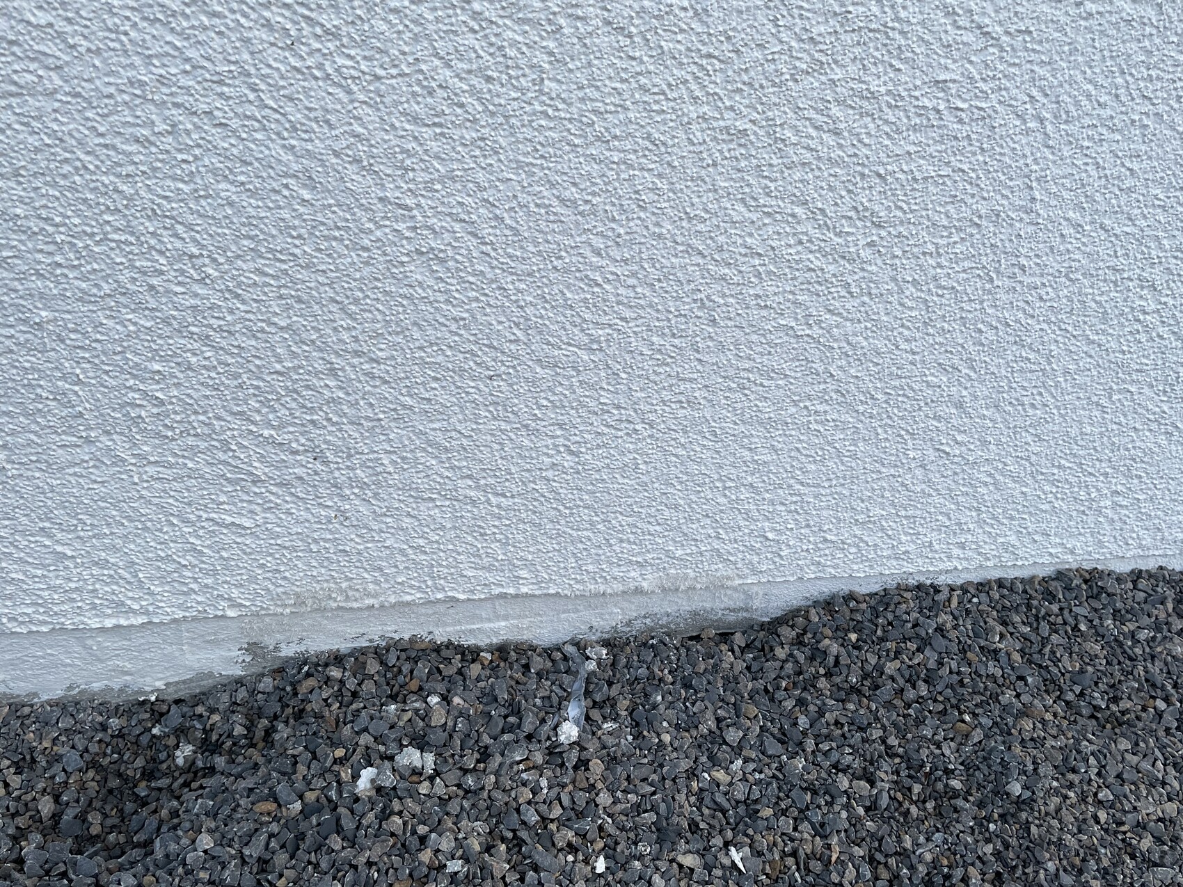 Außenwand mit rauem weißen Putz, untere Betonkante, davor dunkler Kiesboden.