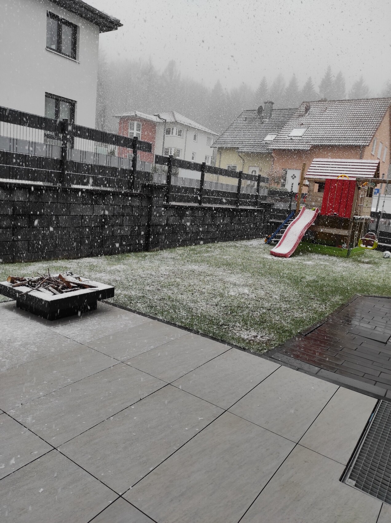 Schneefalliger Hinterhof mit Spielturm und Rutsche, Zaun und benachbarten Häusern.