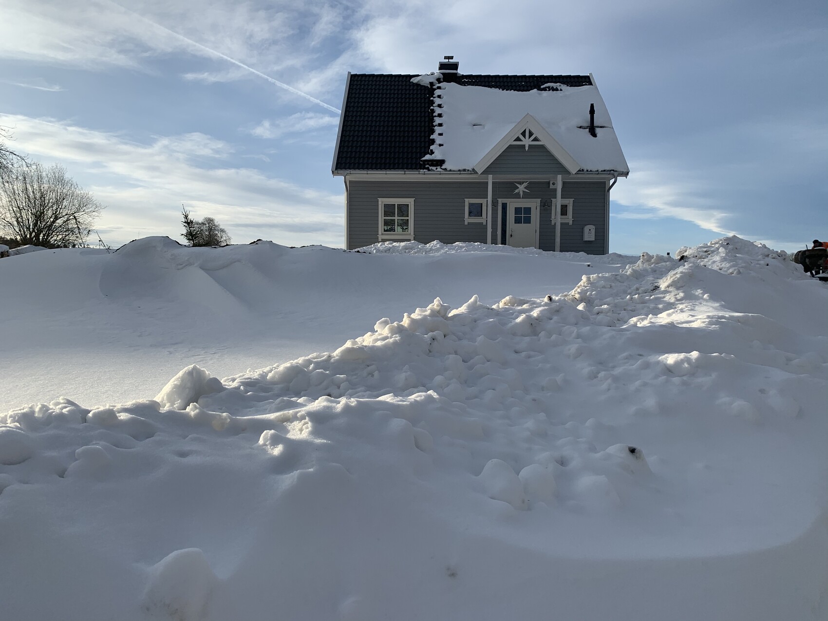 Kleines graues Haus mit verschneitem Dach, von hohen Schneehügeln umgeben, unter blauem Himmel.