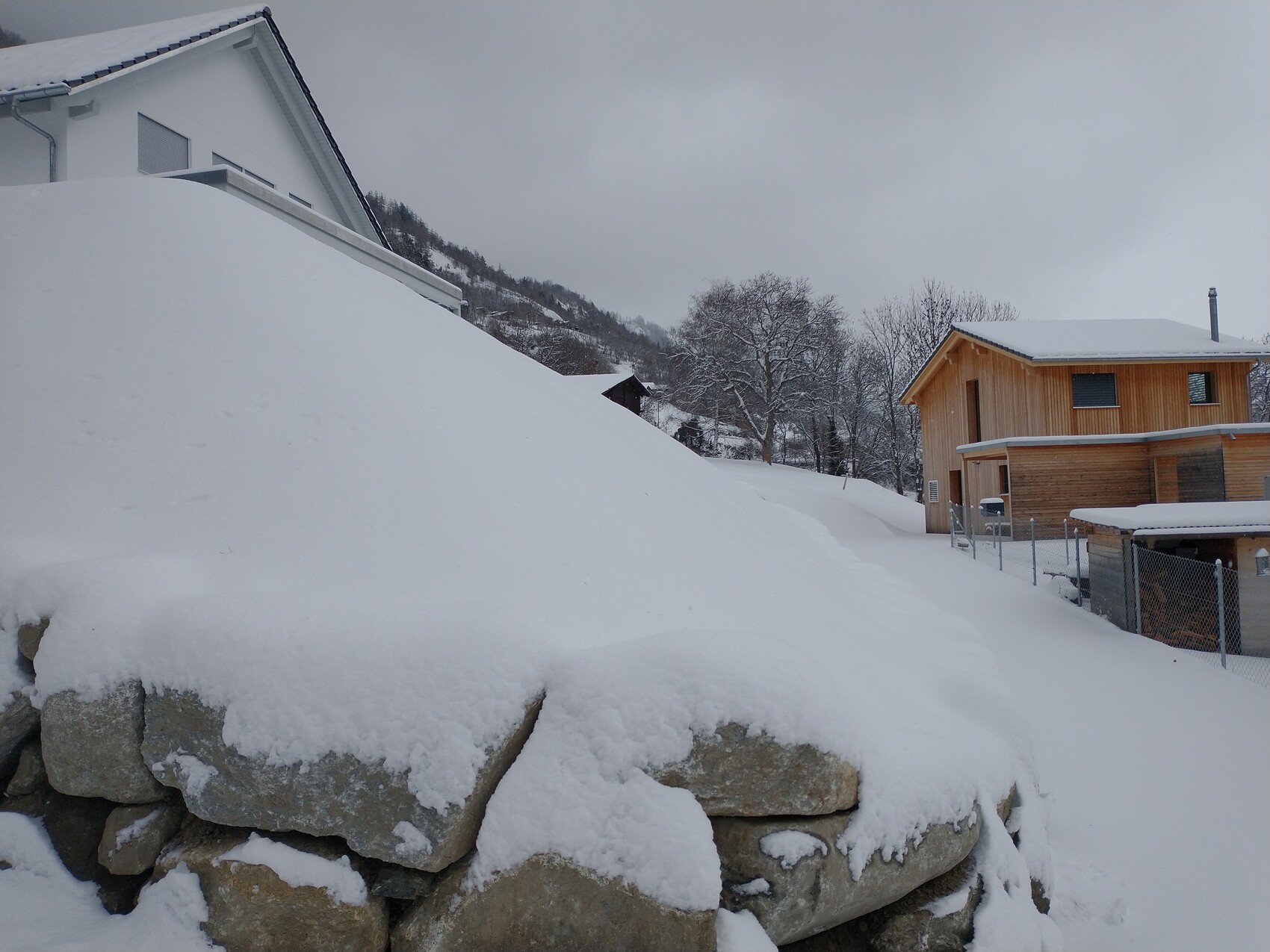 Schneebedeckte Vorstadt: links großes weißes Haus, rechts Holzhäuschen, Steinmauer im Vordergrund.