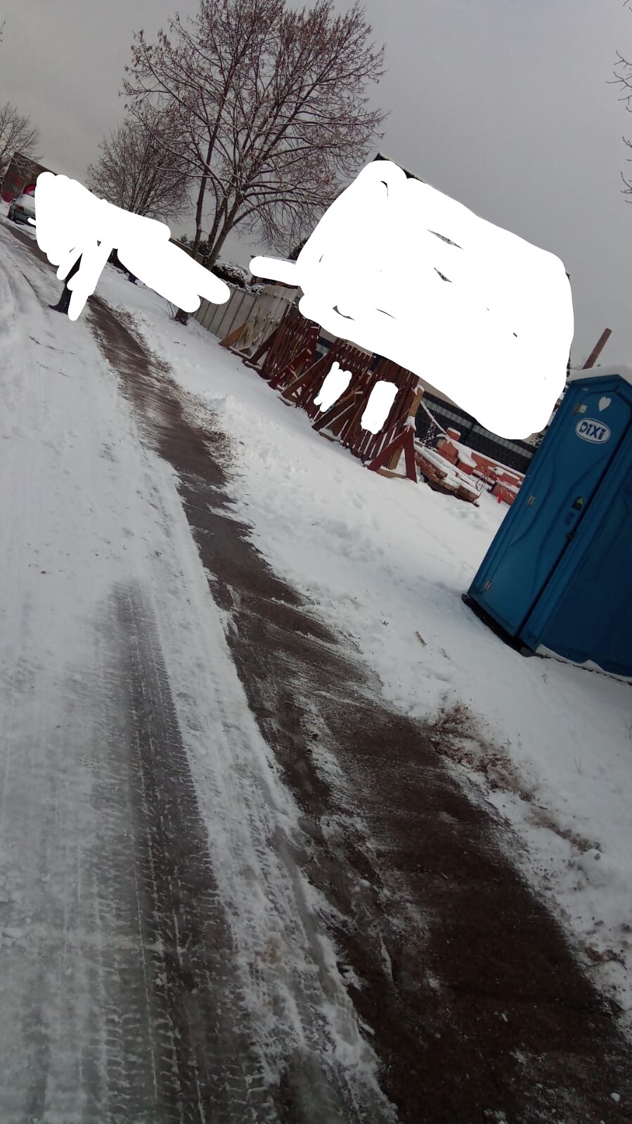 Schneebedeckte Straße mit Gehweg, Bauzaun und blauer Porta-Potty rechts.