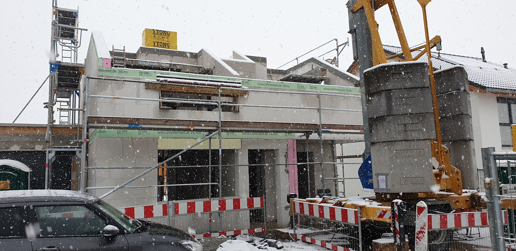 Baustelle im Schnee: Gerüst um zweistöckiges Gebäude, Kran rechts, Absperrband, Auto im Vordergrund.