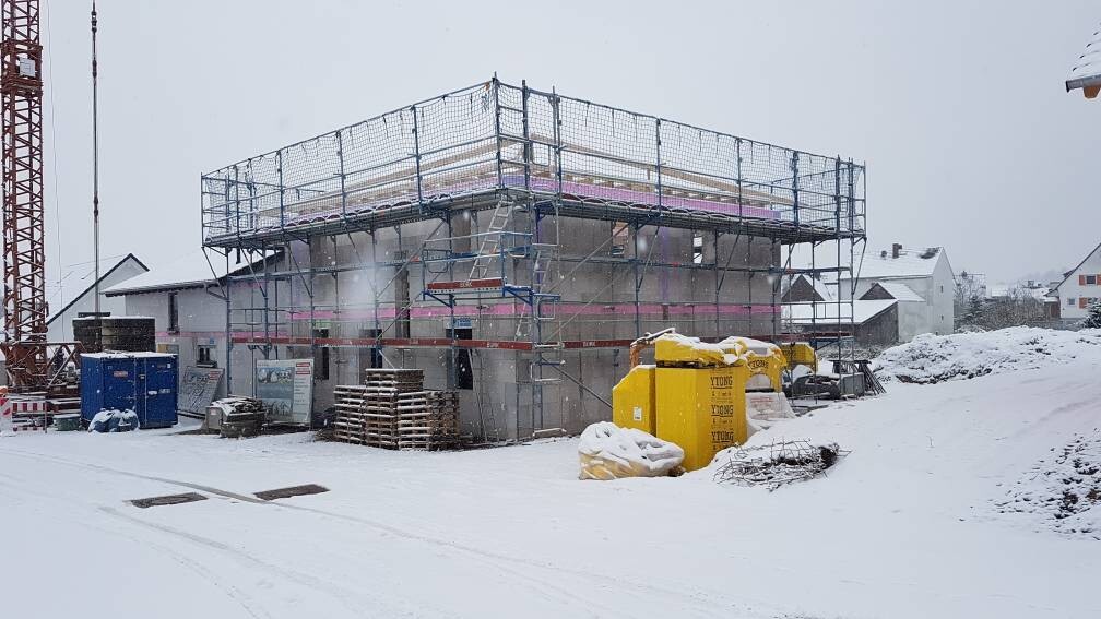 Schneebedeckte Baustelle mit Gerüst, Baucontainern, Kran links und Paletten.