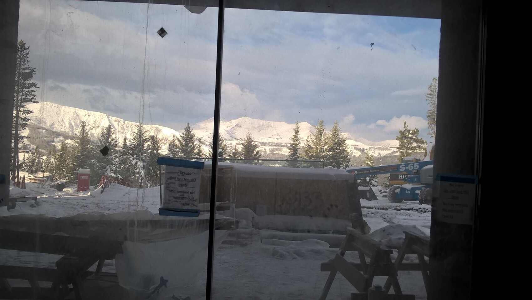 Winterliche Baustelle im Schnee mit Bergen und Tannen; blaue Hebebühne außerhalb des Fensters.
