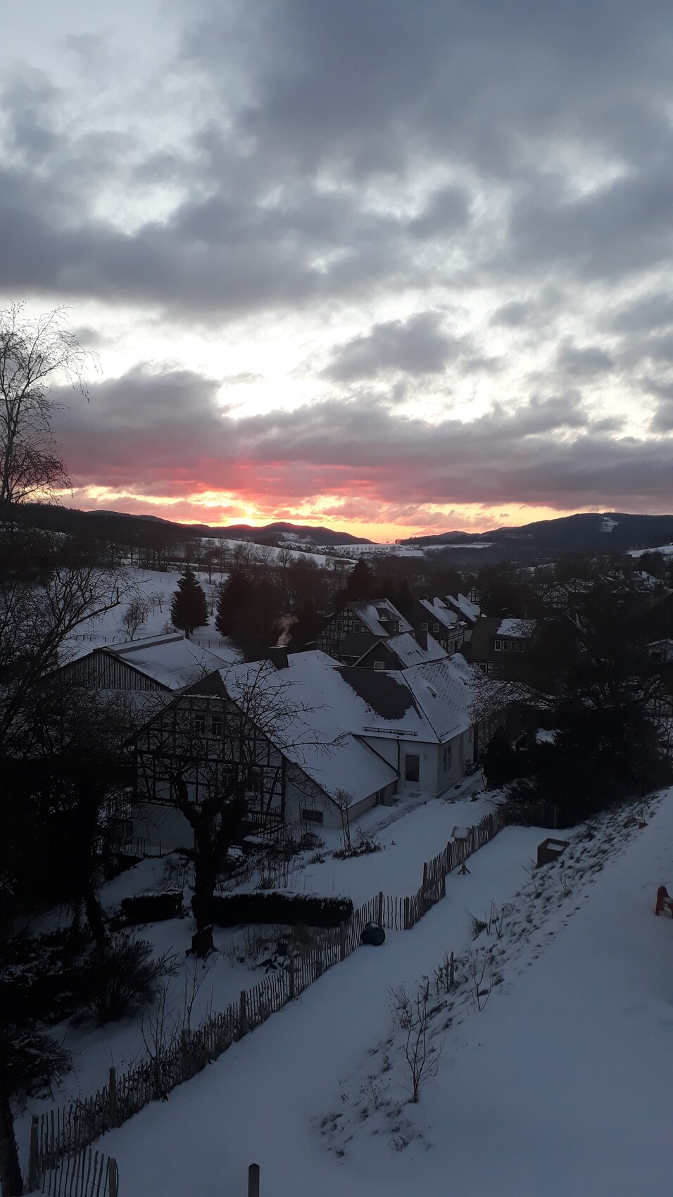 Abendrot über verschneitem Dorf mit schneebedeckten Dächern, Bäumen und Hügeln.