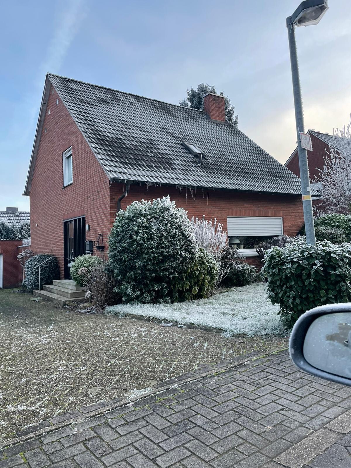 Rotes Einfamilienhaus mit verschneitem Vorgarten, frostige Büsche, Laterne und Autospiegel im Bild.