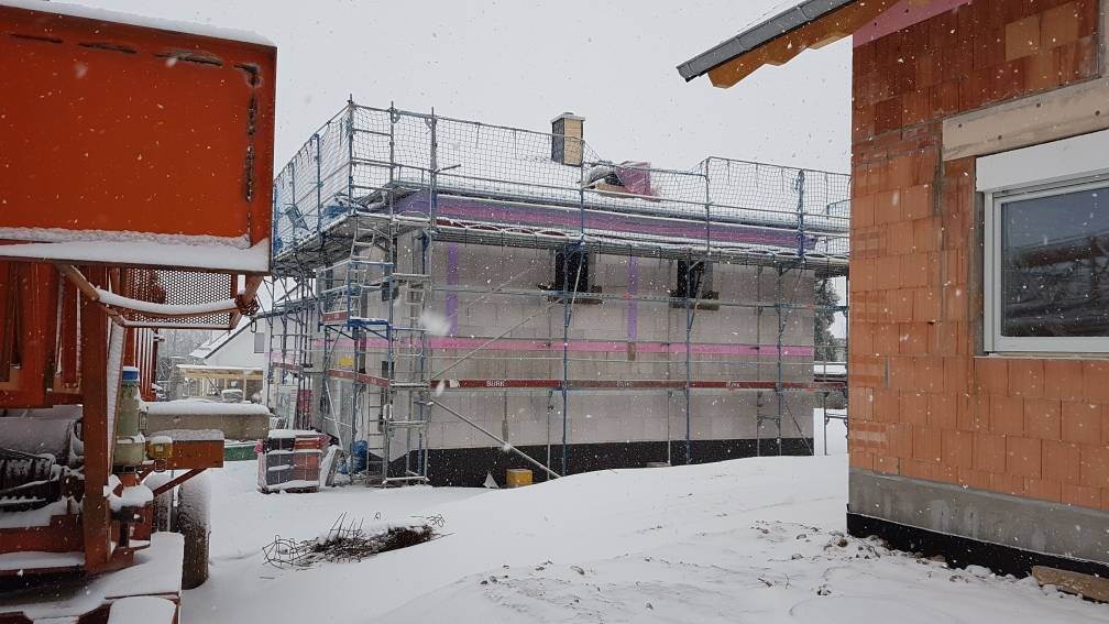 Winterliche Baustelle: Gerüst um ein Rohbauhaus, Schnee fällt, daneben ein orangefarbenes Gebäude.