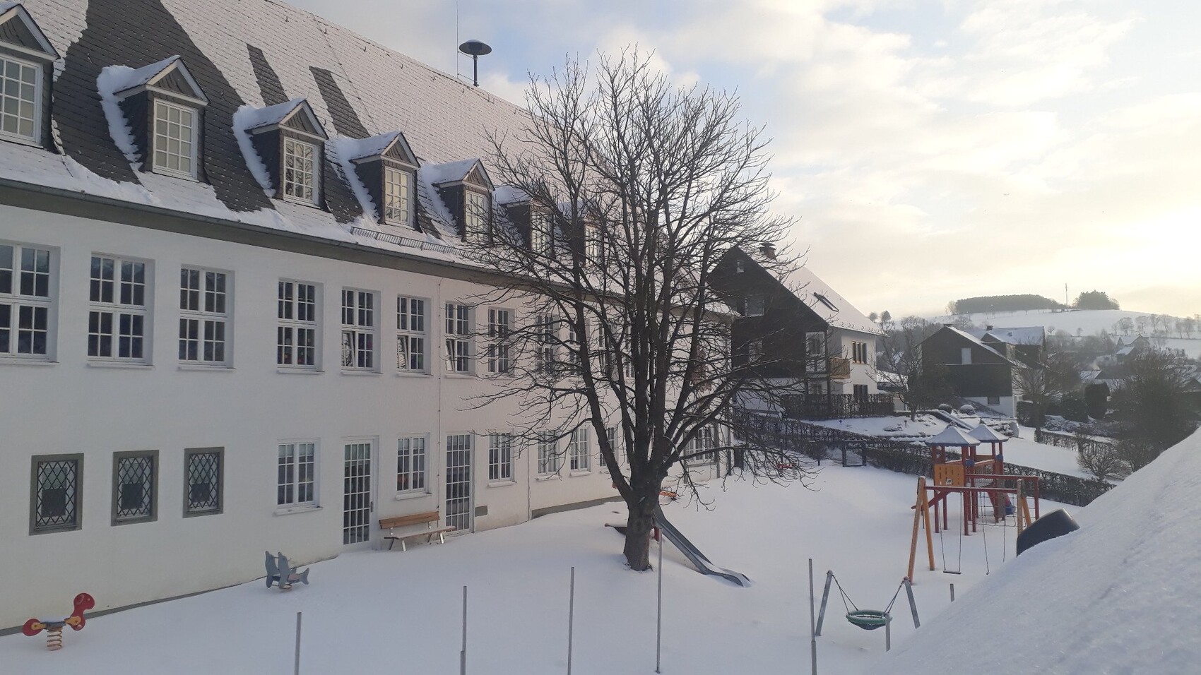 Winterliche Schulhof-Szene: weißes Gebäude, Baum und Spielplatz im Schnee