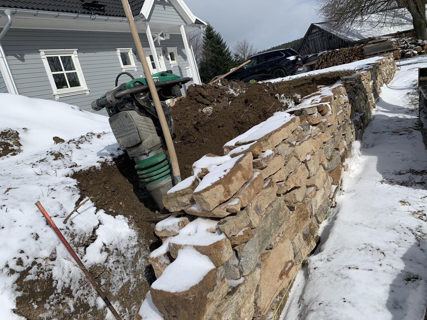 Bau einer Steinmauer am Hang neben einem Haus; Schlamm, Schnee und Pumpe im Vordergrund.