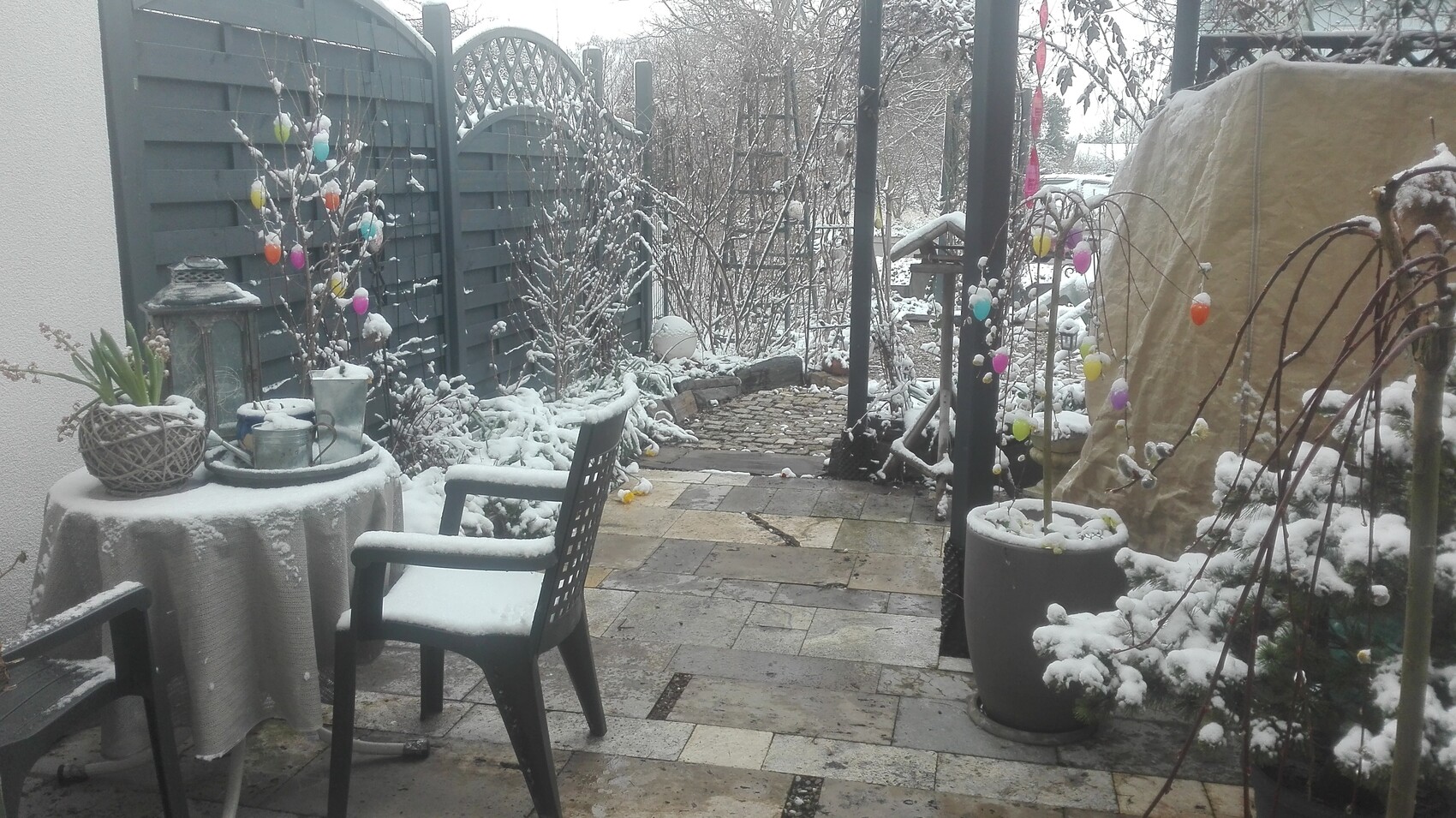 Winterlicher Garten im Schnee mit bunten Eiergirlanden an Sträuchern, Tisch, Stuhl und Pflasterweg.