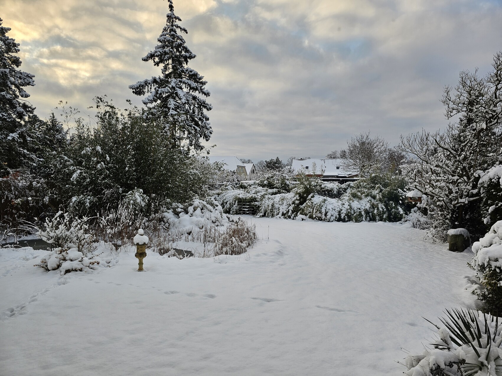 Schneebedeckter Garten mit Bäumen, Sträuchern, Teich und einer Gartenfigur im Vordergrund.