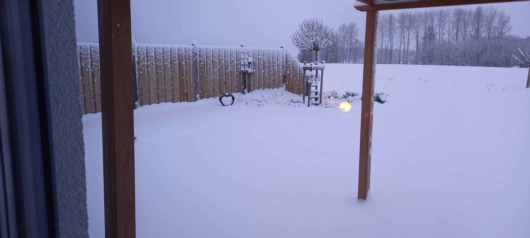 Schneebedeckter Garten mit Holzzaun, Pergola-Stützen und leuchtender Kugellampe im Schnee.