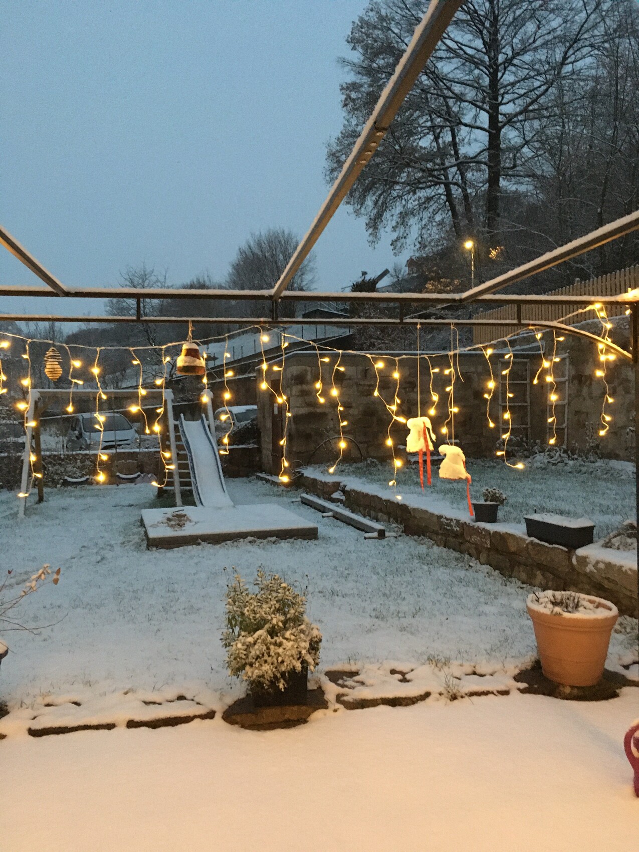 Winterlicher Garten mit Schnee, Lichterketten und Spielgerät unter einer Metall-Pergola
