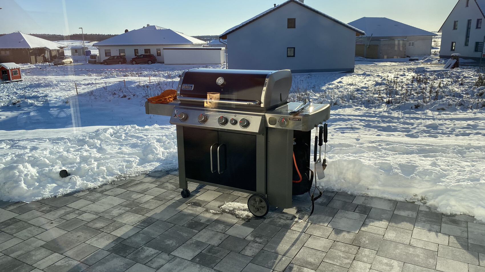 Gasgrill auf gepflasterter Terrasse im Schnee; seitlich hängen Grillutensilien.