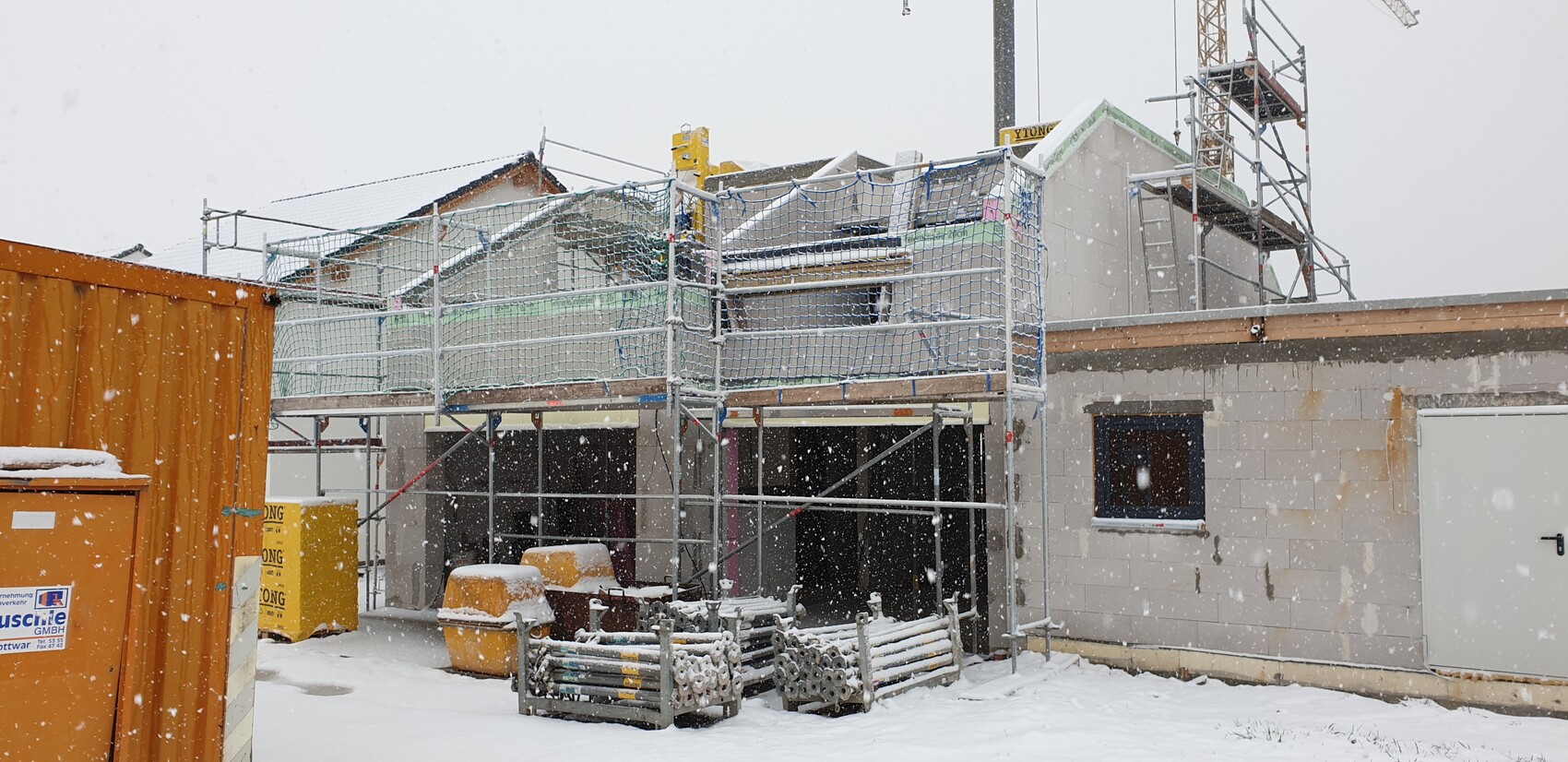 Baustelle im Schnee: Gerüst um ein halbfertiges Haus, Baucontainer links, Kran im Hintergrund.