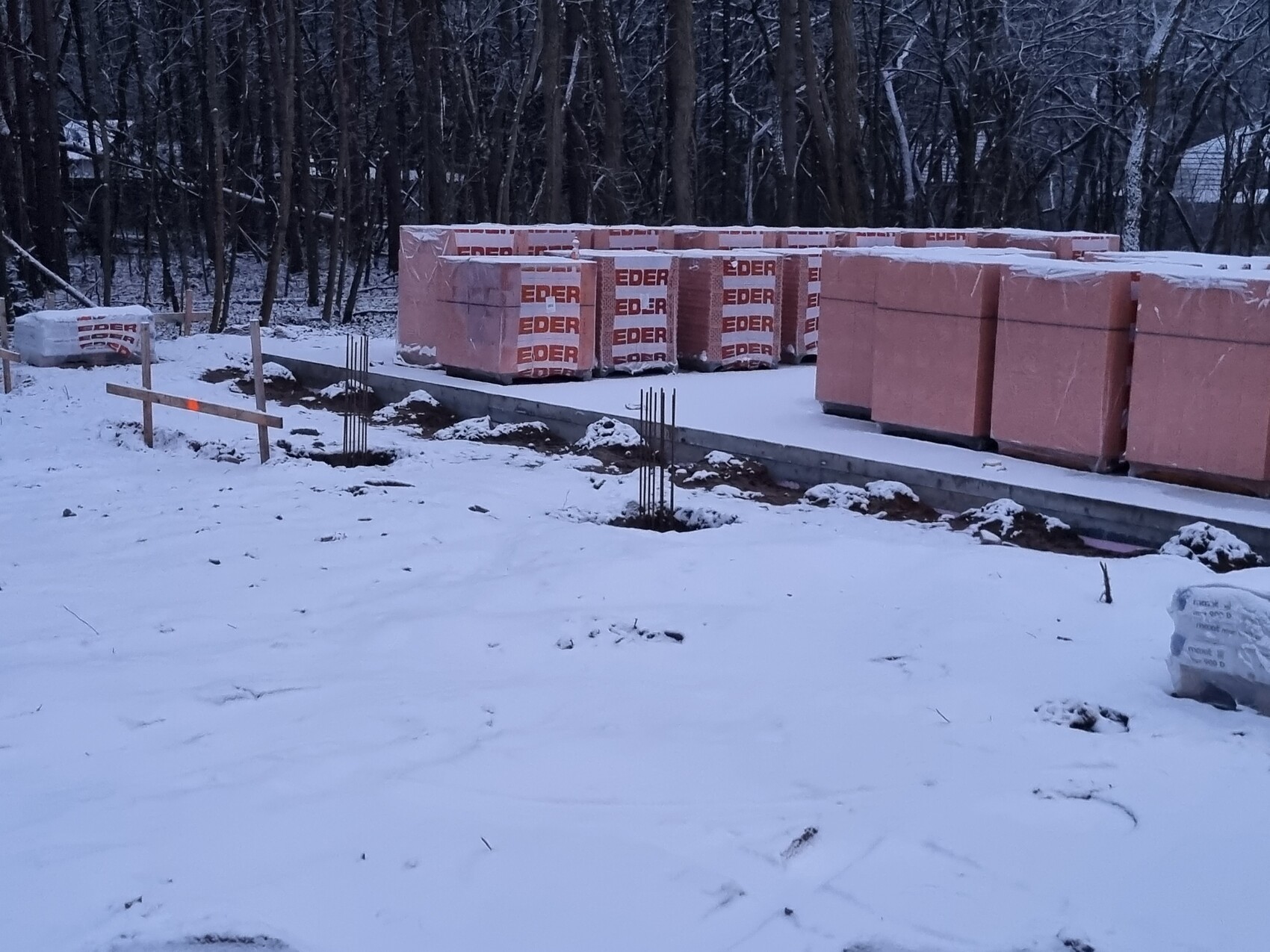 Baustelle im Schnee: Paletten mit Dämmplatten, Bewehrung und Betonfundament sichtbar.