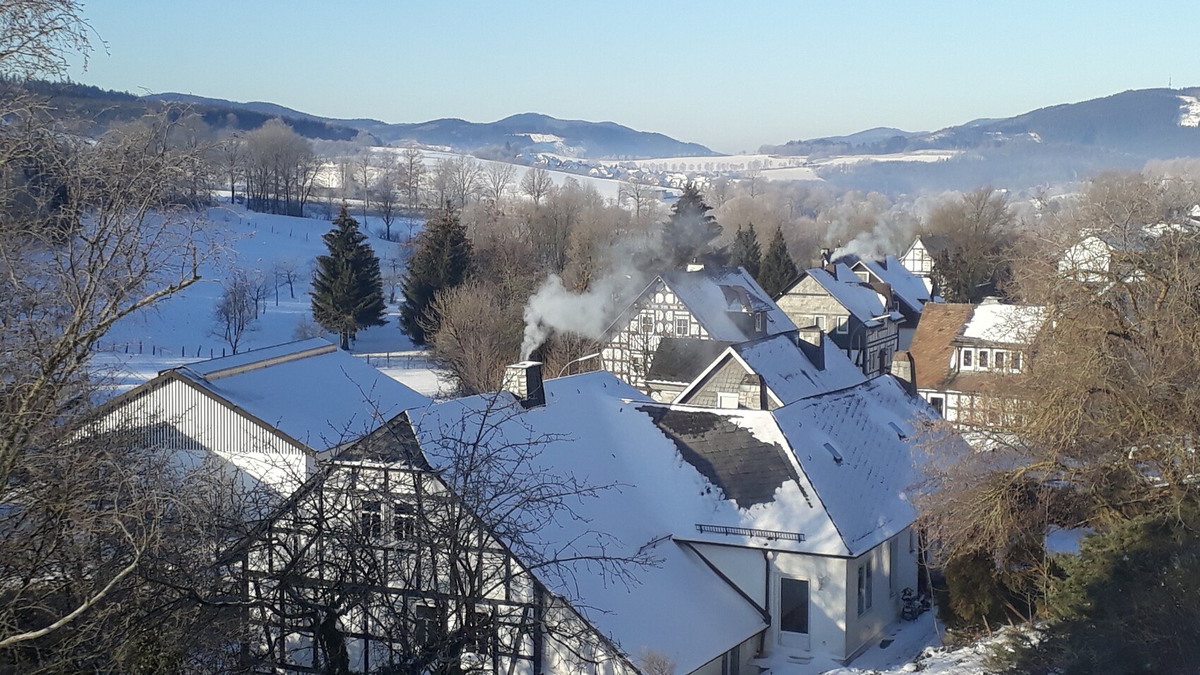 Verschneites Dorf mit Fachwerkhäusern, Rauch aus Schornstein, umgeben von Winterlandschaft.