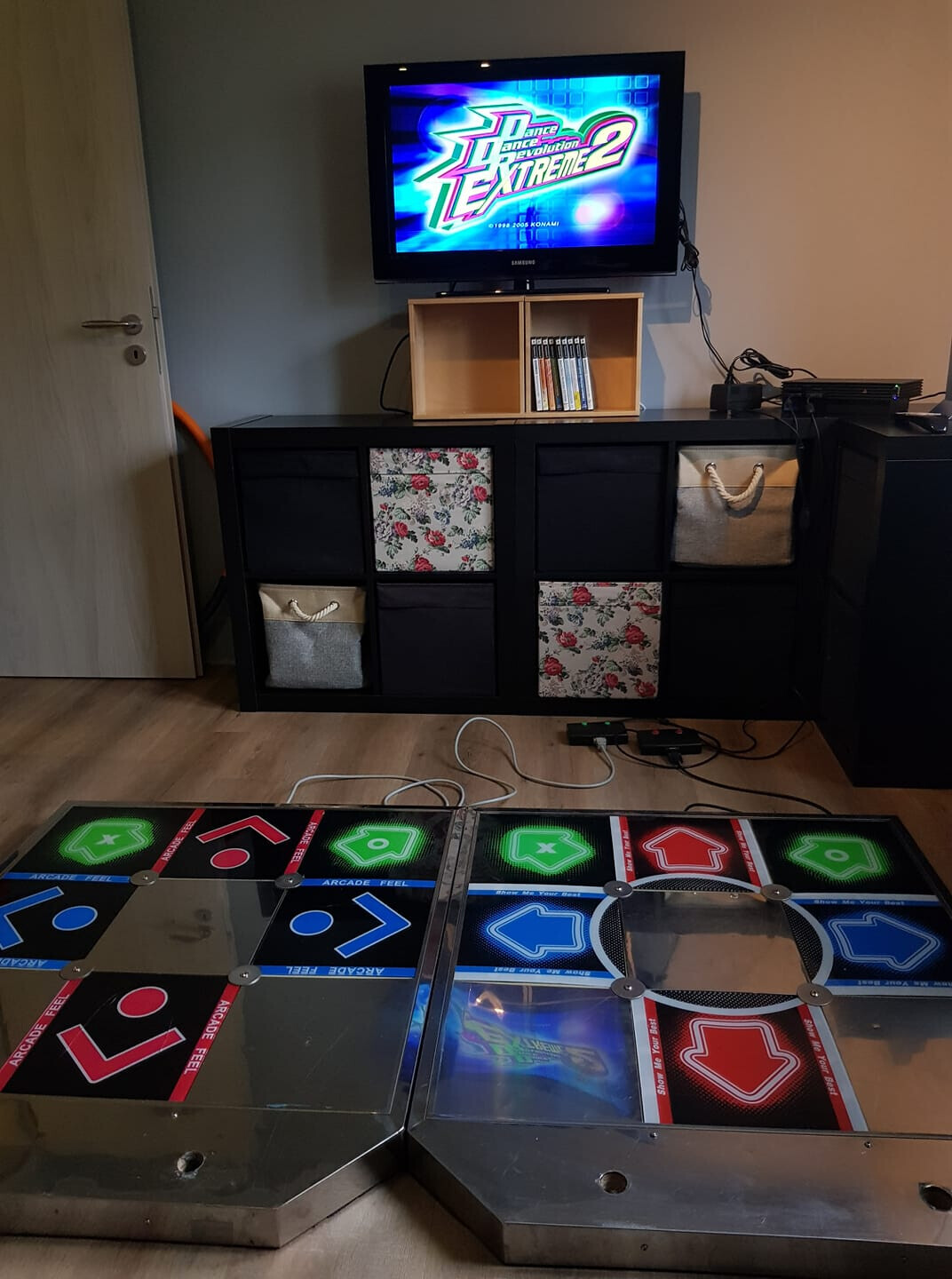 Wohnzimmer mit Fernseher auf Regal, Spielkonsole und zwei großen Dance-Mats auf dem Boden.