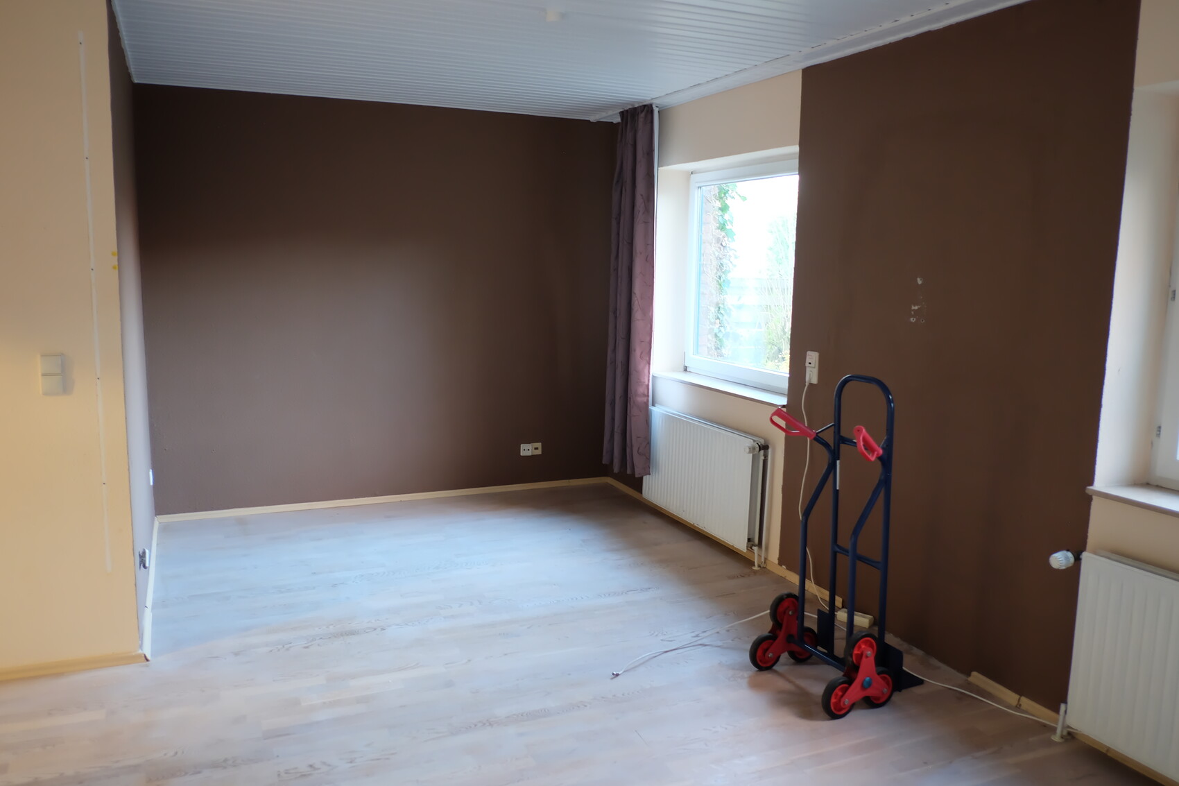 Leerer Raum mit braunen Wänden, Fenster, Heizkörper und blaue Handkarre.