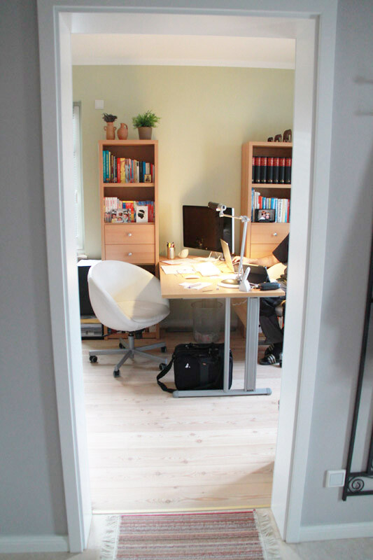 Büro durch Tür: Schreibtisch mit Monitor, Lampe, Regale voller Bücher, Pflanze, weißer Stuhl.