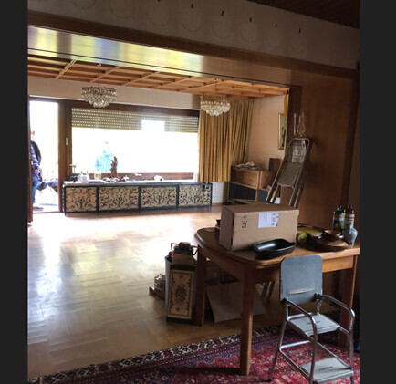 Wohnzimmer mit Esstisch, Stuhl, Kronleuchter und Blick auf Sofa vor großen Fenstern