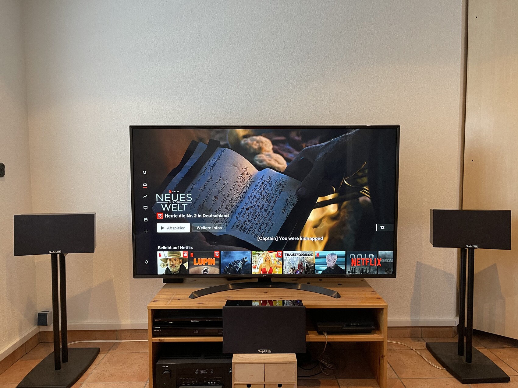 Flachbildfernseher auf Holztisch, flankiert von zwei Standlautsprechern; Netflix-Oberfläche sichtbar.